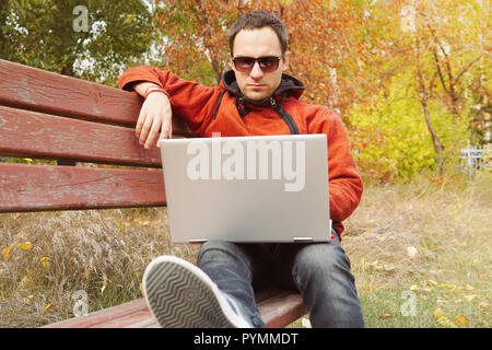 Freelancer programmer sitting at laptop on a bench in Park outdoors.. Young man working on his laptop connect to wireless internet in rural area. Inte Stock Photo