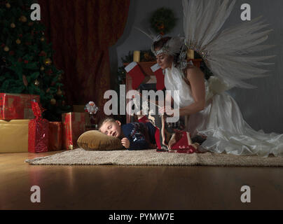 Sleeping boy under the Christmas tree and guardian angel covering him with a blanket. Angel in white dress with natural feathers. Stock Photo