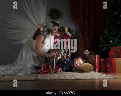 Sleeping boy under the Christmas tree and guardian angel covering him with a blanket. Angel in white dress with natural feathers. Stock Photo