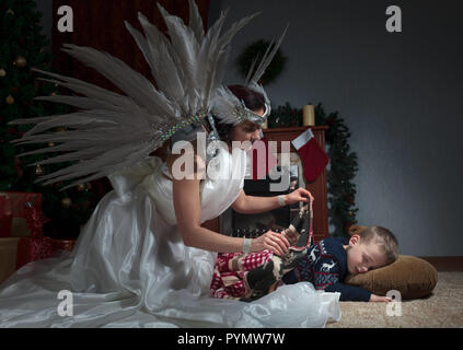 Sleeping boy under the Christmas tree and guardian angel covering him with a blanket. Angel in white dress with natural feathers. Stock Photo