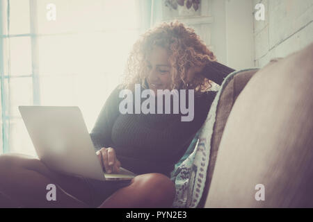 Curly woman with gray sweater sitting on brown sofa and blue blankets relaxes looking at the computer on an autumn day. Technology concept Stock Photo