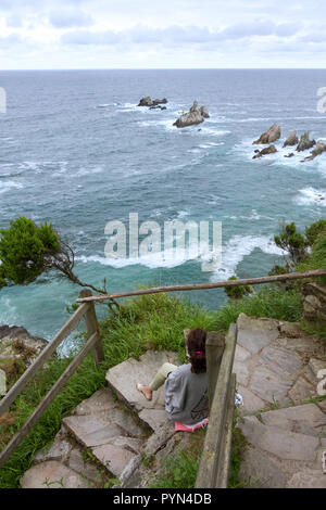 Gueirua beach, Asturias, Spain. General view of tourist woman watching the rough sea breaking in the reef sat on a flight of stone stairs. Stock Photo