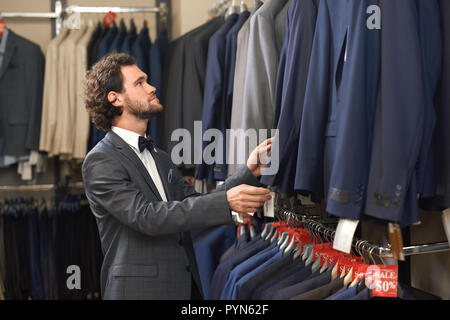 Young handsome man in costume looking for jacket in shop. Brunnete curly haired guy buying clothes in fashion boutique. Customer in store touching clothes on hangers. Concept of fashion and shooping. Stock Photo