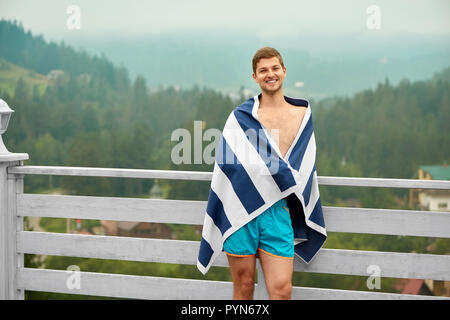 Happy smiling guy, wearing in blue shorts, and wrapped in striped towel, standing on wooden terrace of mountain resort. Young man, enjoying rest, leaning on railing, smiling and looking at camera. Stock Photo