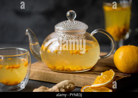 Glass teapot with sea buck thorn tea with ginger at wooden board on black background. Orange and ginger near Stock Photo