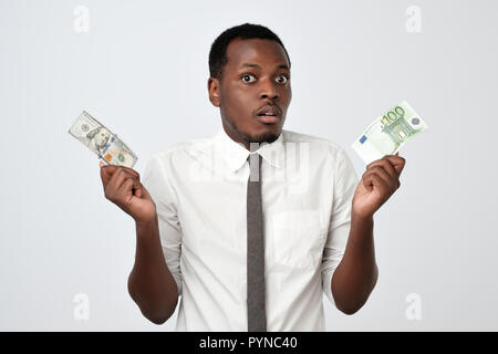 Young attractive african man holding USA currency and euro currency deciding which one to choose Stock Photo