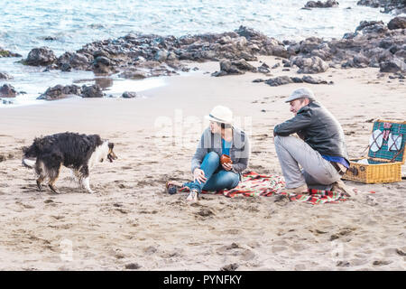 Alternative family with one lady a man and a dog together at the beach enjoying a picnic in friendship and partenership. Having fun in outdoor leisure Stock Photo
