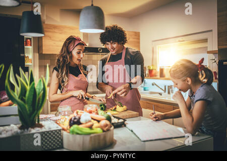 Happy parents and their daughter cooking together in the kitchen while little girl doing her homework on the kitchen table. Stock Photo