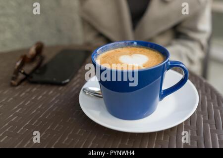 Blue cup with cappuccino, on a table in street cafe, drawing a heart. Stock Photo