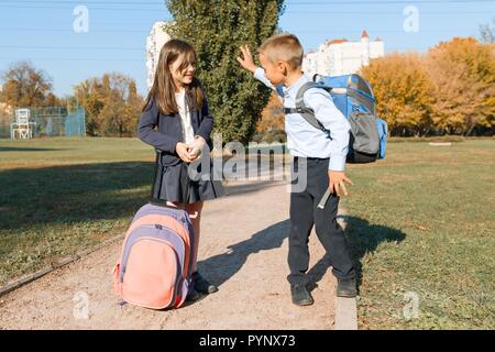 Boy and girl, primary school students with backpacks go to school. Sunny day background, road in the park. Stock Photo