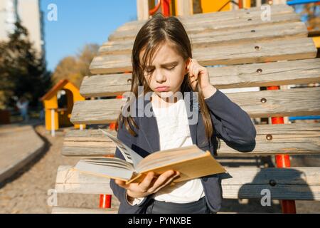 Outdoor portrait of offended little girl. A girl is reading thick book, offendedly pouting her lips. Stock Photo