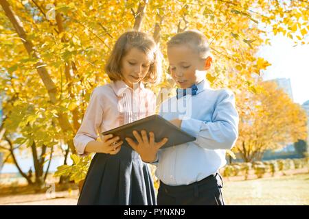 Two kids watching digital tablet, background autumn sunny park, golden hour. Stock Photo