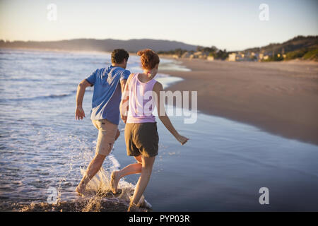 Young couple holding hands while running through water on a sandy beach. Stock Photo