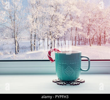 Winter background - cup with candy cane on windowsill and winter forest outdoors. Cozy still life with concept of spending winter indoors with Christm Stock Photo