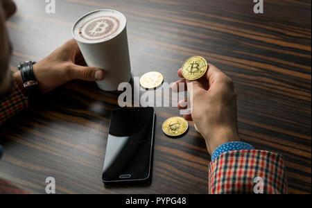 Rich man holding a bitcoin coin and drinking a coffee with a phone and gold coins of bitcoin on a table. Profit from mining of bitcoins. Stock Photo