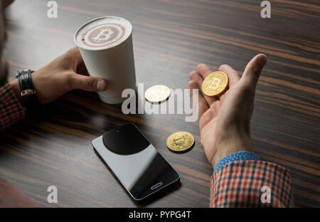 Rich man holding a bitcoin coin and drinking a coffee with a phone and gold coins of bitcoin on a table. Profit from mining of bitcoins. Stock Photo