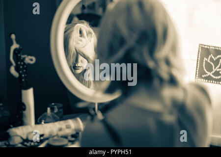 Cinematic portrait of woman sitting on front of mirror in her bedroom and looking troubled.Shot from behind with reflection of hand covering face show Stock Photo