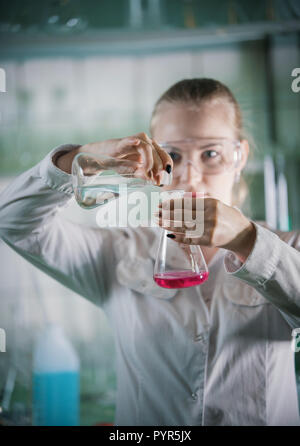 Chemical laboratory. Young blonde woman holding two flasks with liquid in it. About to transfuse the liquid Stock Photo