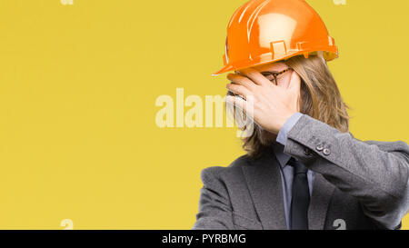 Young handsome architec man with long hair wearing safety helmet over isolated background covering eyes with hands and doing stop gesture with sad and Stock Photo