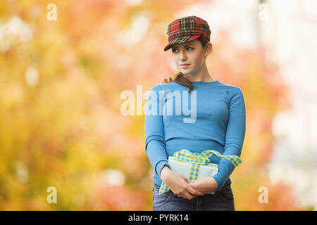 Young woman in a plaid cap holding a wrapped gift tied with a bow outside. Stock Photo
