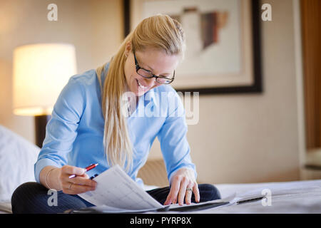 Young businesswoman smiles as she checks a personal organiser while she sits cross-legged on a bed. Stock Photo