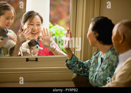 Young girl and her mother hold a puppy each and smile and wave through a window to a senior couple sitting inside. Stock Photo