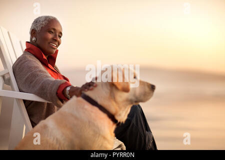 Smiling senior woman petting her dog while sitting on a beach. Stock Photo