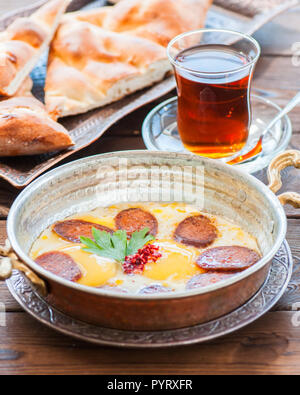 Turkish breakfast  - fried eggs with sausages (sucuk) and spices in a pan, glass of tea on a wooden background. Stock Photo