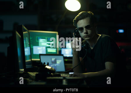 Hacker with sunglasses holding a telephone receiver and computer screens, coding and system hacking concept Stock Photo