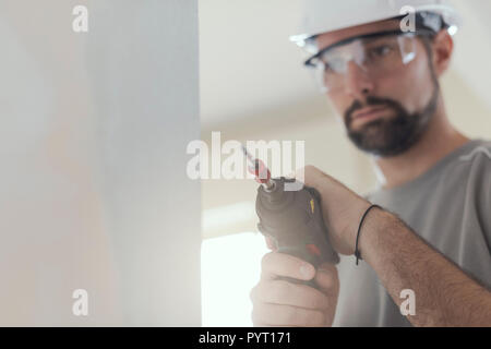Professional construction worker using a drill, he is wearing a safety helmet and goggles Stock Photo