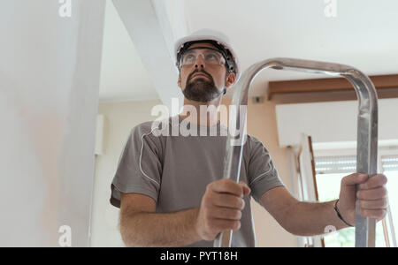 Professional construction worker doing a home makeover, he is standing on a ladder and looking around Stock Photo