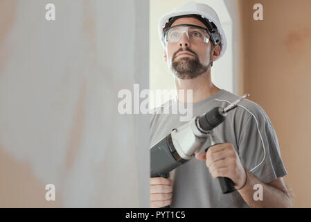Professional construction worker using a drill, he is wearing a safety helmet and goggles Stock Photo