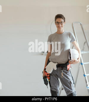 Professional repairwoman with tool belt and safety helmet doing a home renovation, she is posing and holding a drill Stock Photo