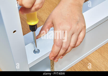 Handyman tightens a nut in a piece of wooden table with a screwdriver, the furniture assembler holds the part of the table with his second hand. Stock Photo