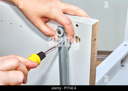 Handyman tightens the bolt fixings in the drawer slide brackets on the surface of a white wooden table with a screwdriver. Stock Photo