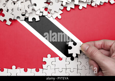 Trinidad and Tobago flag  is depicted on a table on which the human hand folds a puzzle of white color. Stock Photo