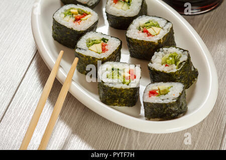 Sushi rolls with rice, pieces of avocado, cucumber, red bell pepper and lettuce leaves on ceramic plate, chopsticks on wooden desk. Vegetarian food Stock Photo