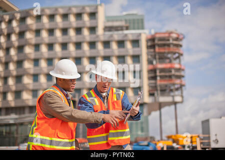 Two engineers in high visibility vests and hardhats discuss building plans on a construction site. Stock Photo