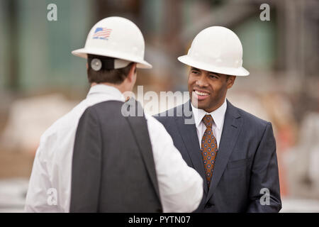 Two young engineers having a business discussion while wearing suits and hardhats. Stock Photo