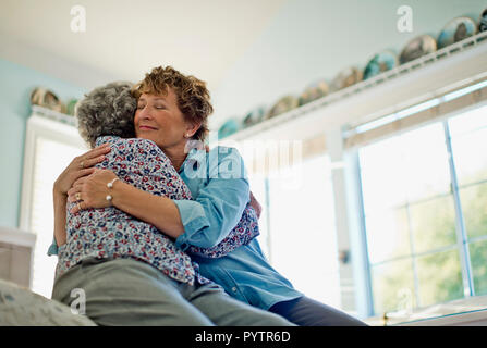Mature woman gently smiles as she embraces her elderly mother as they sit on the edge of a comfortable bed. Stock Photo
