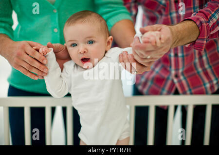 Cute smiling baby boy is supported by his parent's hands as they help him stand in his cot for the first time. Stock Photo