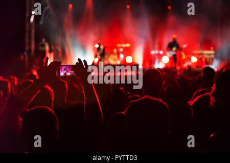 hands filming concert with smartphone, crowd of spectators watching musicians playing music and singing on stage Stock Photo