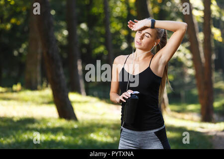 Sportish long-haired young woman wearing black shirt resting before the jogging and drinking water in the sunny park, healthy lifestyle and people concept Stock Photo