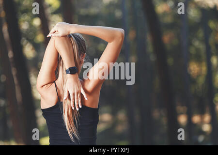 Portrait of young athletic long haired woman wearing sportish clothes stretching her arms and listening music in the sunny park, healthy lifestyle and people concept Stock Photo