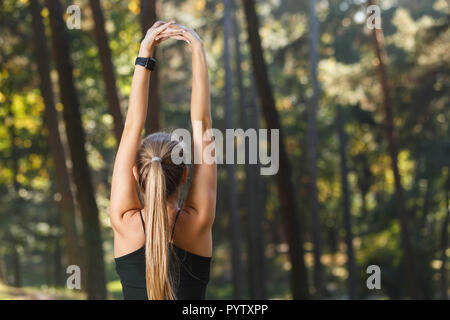 Portrait of young athletic long haired woman with raised arms wearing sportish clothes stretching her arms and listening music in the sunny park, healthy lifestyle and people concept Stock Photo