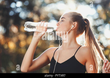Portrait of young athletic long haired woman wearing sportish clothes listening music, touches her hair and drinking water in the sunny park, healthy lifestyle and people concept Stock Photo
