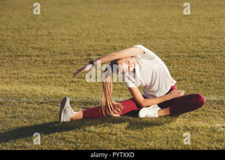 Fitness positive blonde long haired woman in sportswear listening music and doing pilates exercises at the football stadium outdoor, healthy lifestyle and people concept Stock Photo