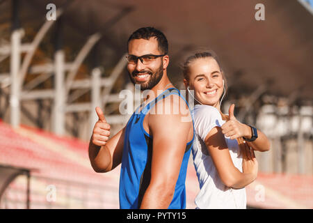 Portrait of multicultural handsome bearded man in blue shirt and sportish pretty blonde long haired woman in white t-shirt standing back to each other and showing thumbs at the football stadium outdoor, healthy lifestyle and people concept Stock Photo