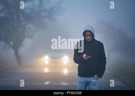 Young man walking on road and using phone. Car approaching in thick fog. Stock Photo