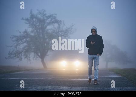 Young man walking on road and using phone. Car approaching in thick fog. Stock Photo